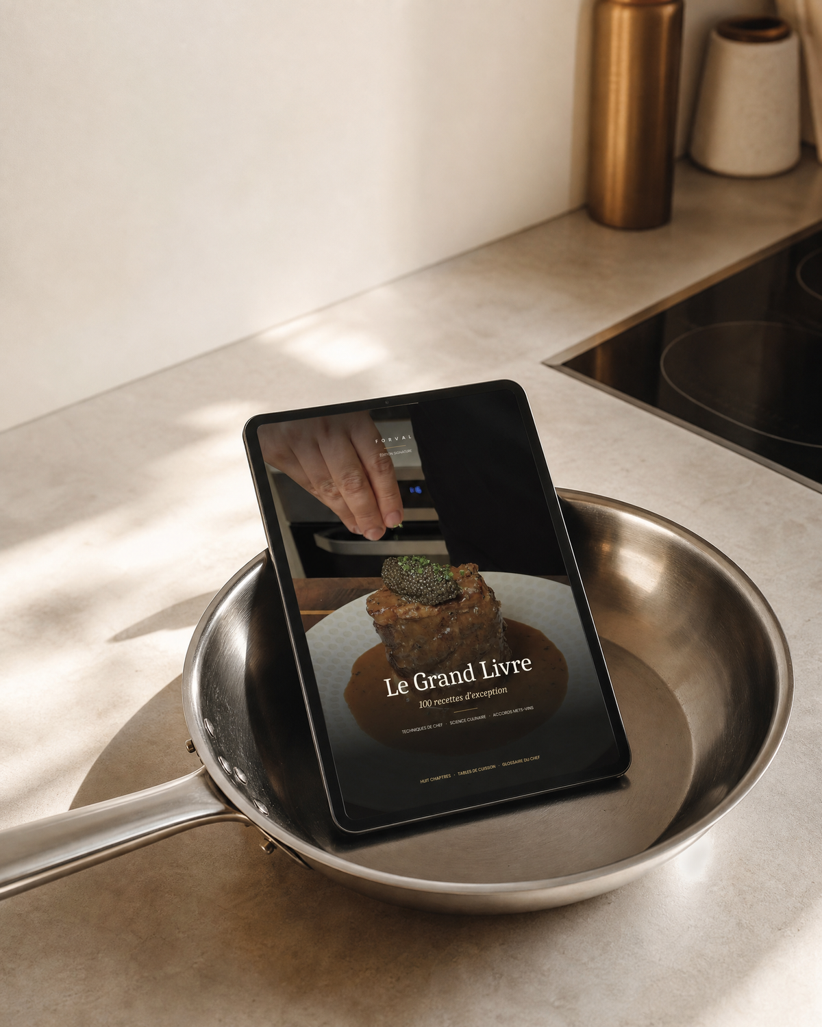 Tablet showing the cover of the Forval Grand Book, leaning in a stainless steel pan on a kitchen worktop.